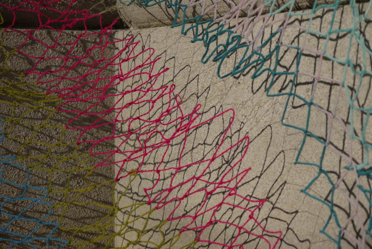close-up of a loose net, made with yarn of a variety of color and thickness. it hangs close to a concrete wall and casts a sharp shadow of its diamond structure.