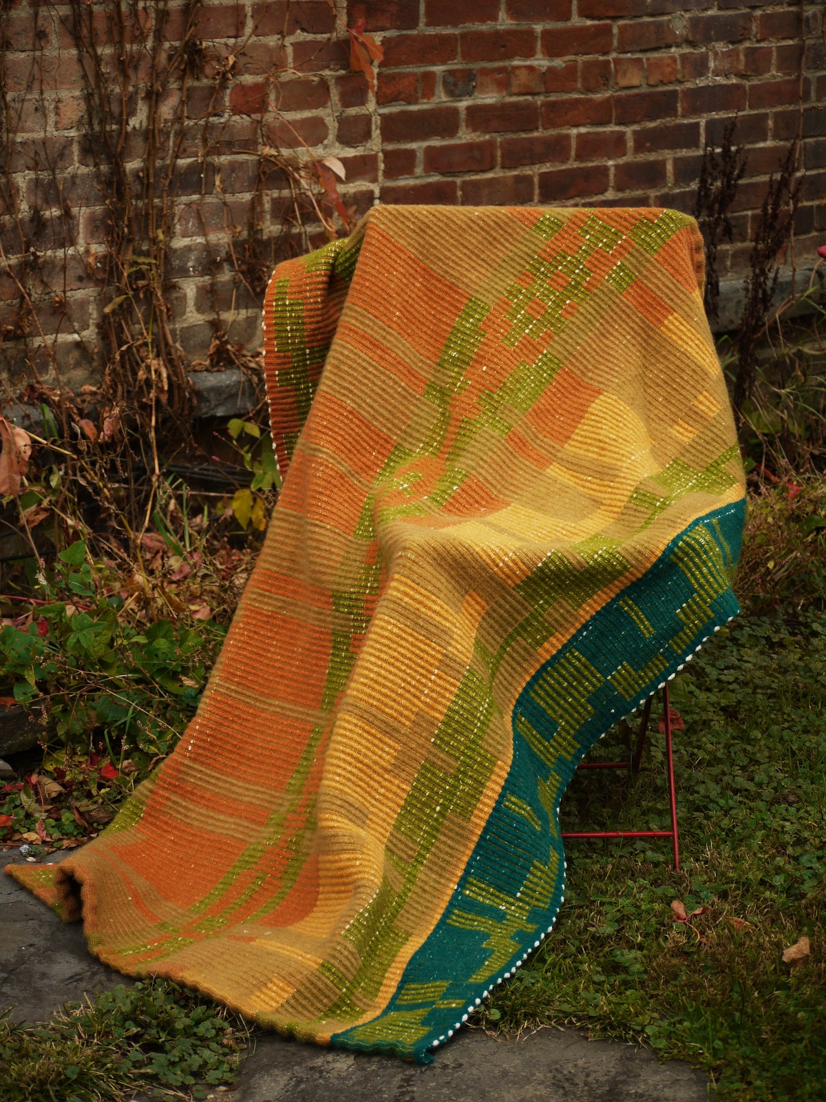 a long rug draped over a chair. it has geometric stripes of green, orange, gold, and blue. behind it, vines climb a brick wall. 