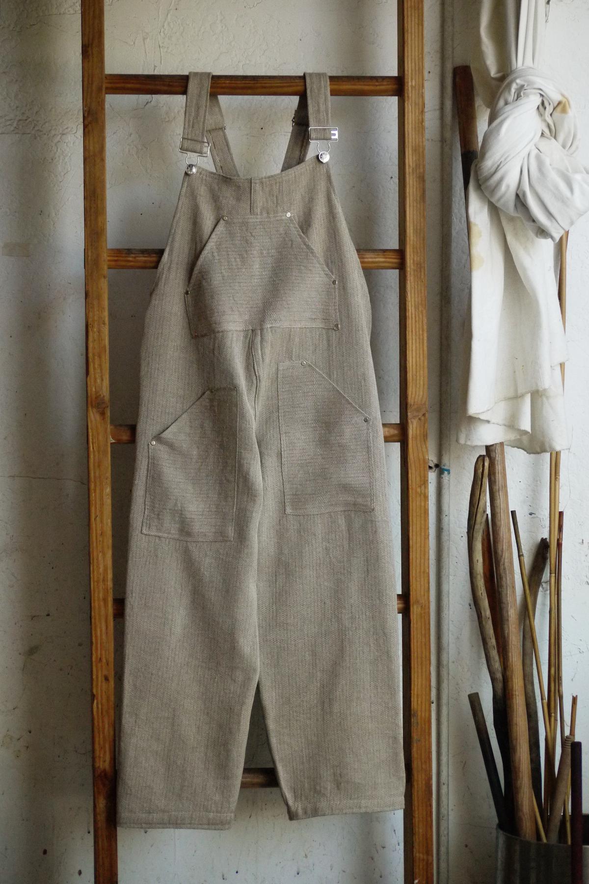 a pair of tan overalls hanging from a wooden ladder. it has boxy dimensions, silver buckles, and large angular pockets.