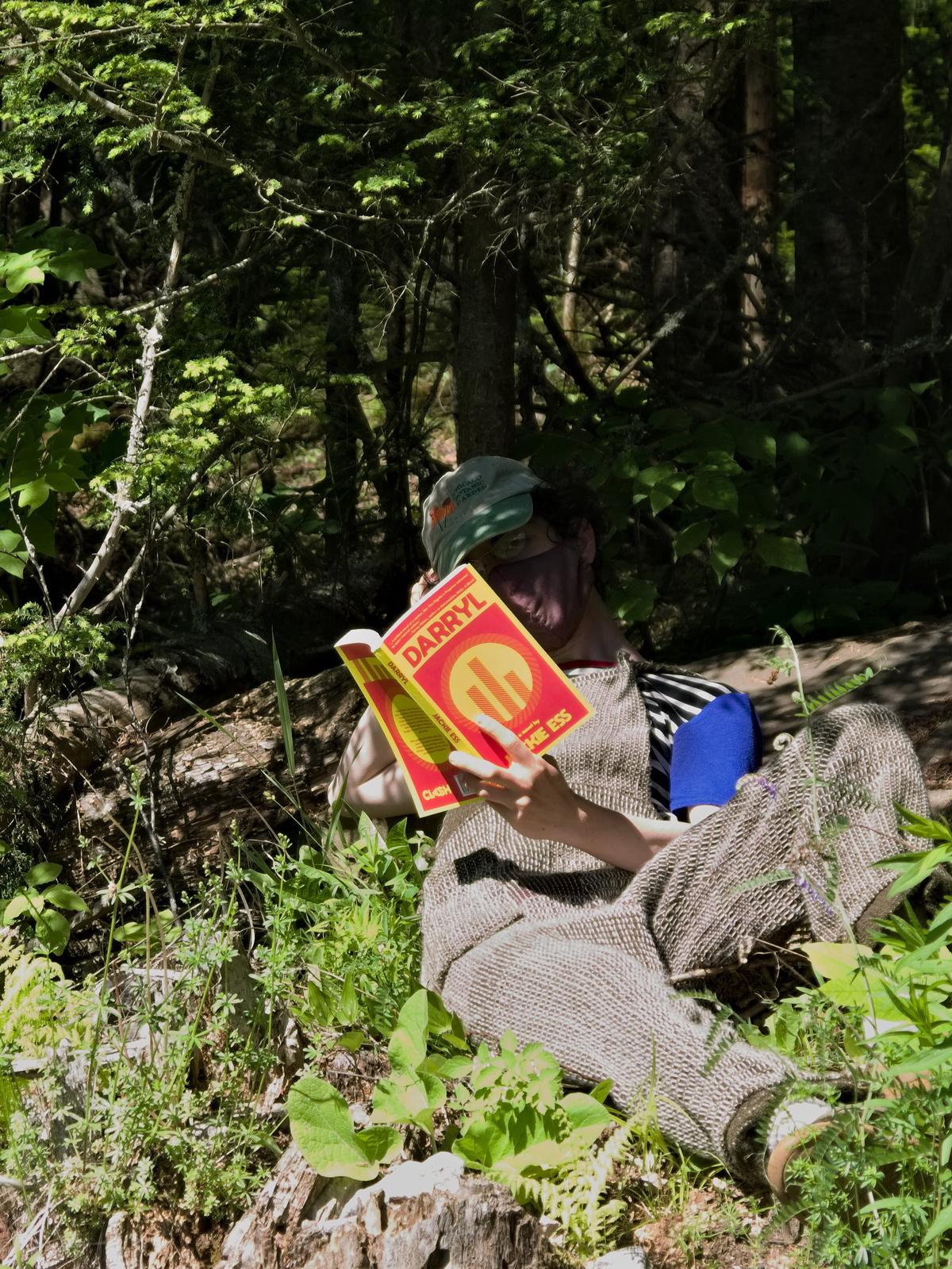 a figure in overalls, reclining in a vibrant forest, with their face in shadow, and reading the novel, Darryl by Jackie Ess.