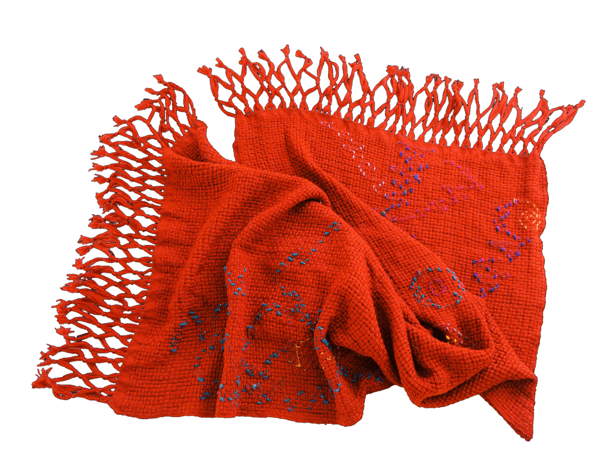 an orange-red shawl, rumpled on a transparent background. it is soft yarn in a loose grid, and inlay of blue, purple, and yellow squiggle over the surface. the fringe is tied in diamonds.