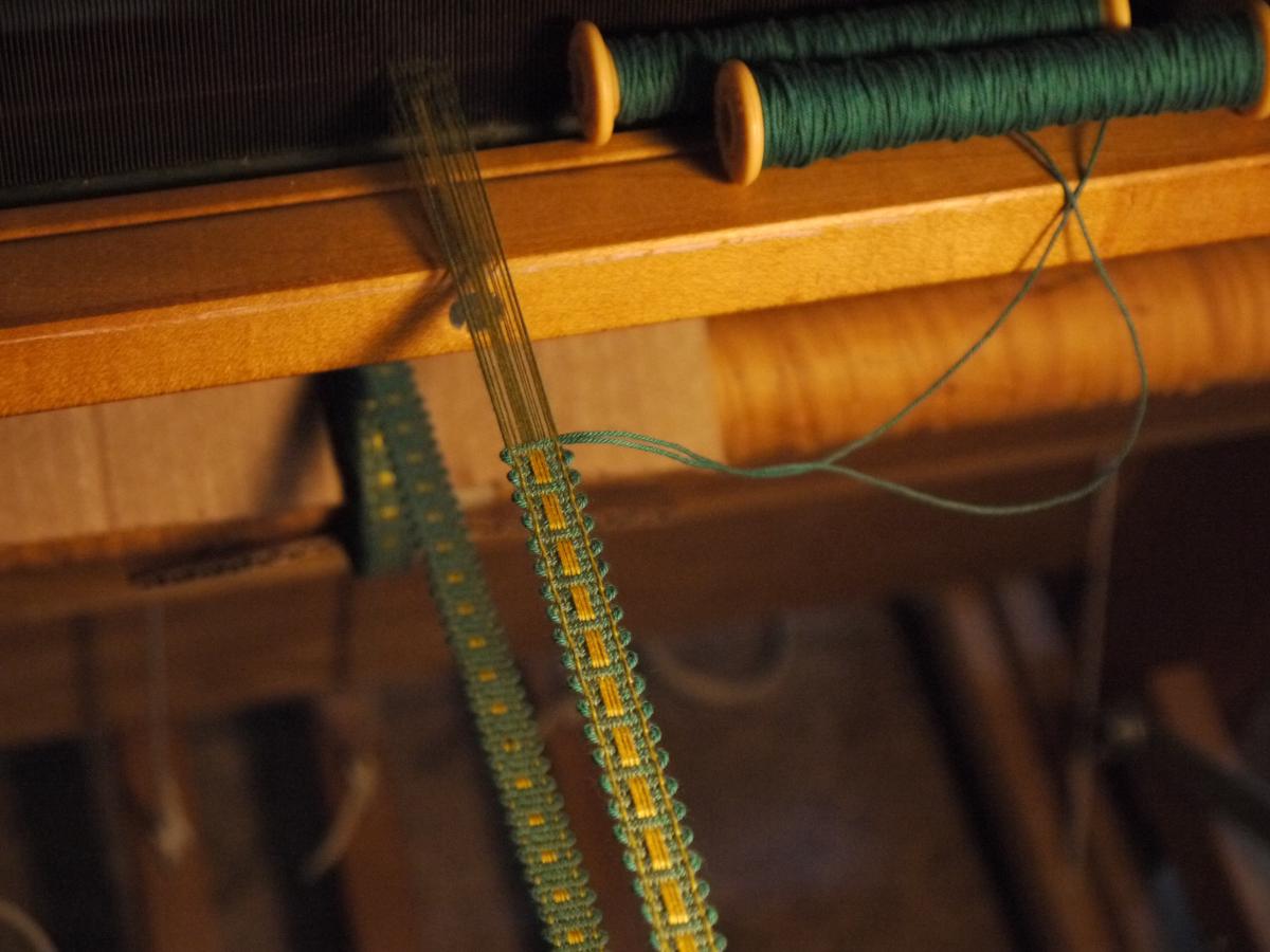 a thin green line of trim, stretched on a loom. two green threads snake out from the top and coil around a pair of bobbins resting on a wooden beam.